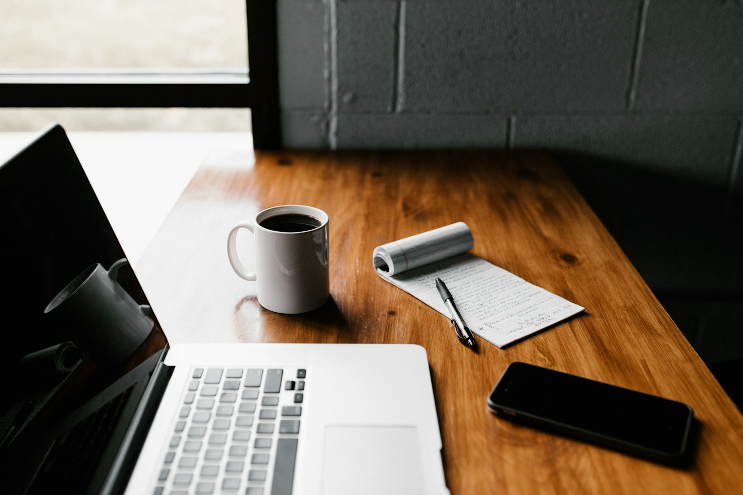 A laptop, a mug of coffee and a notepad on a wooden desk